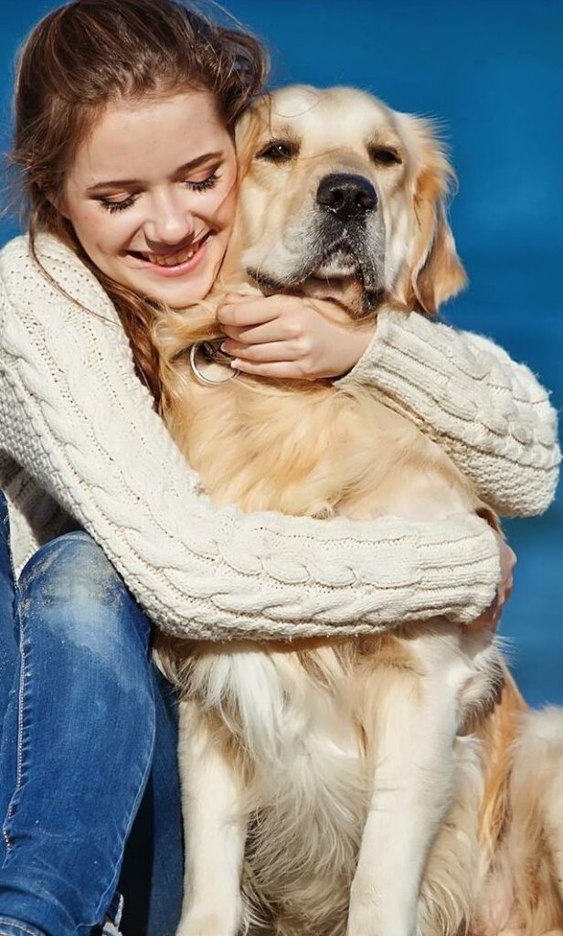 Jeune femme souriante en pull torsadé blanc serrant tendrement dans ses bras un golden retriever, tous deux profitant d’un moment complice en extérieur sous un ciel bleu.