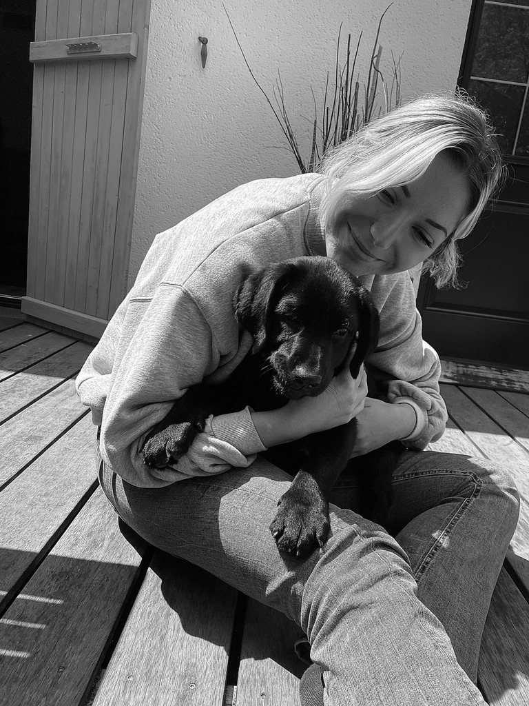 Femme assise sur une terrasse en bois, souriante et câlinant un chiot noir dans ses bras, capturée en noir et blanc dans un moment de tendresse.
